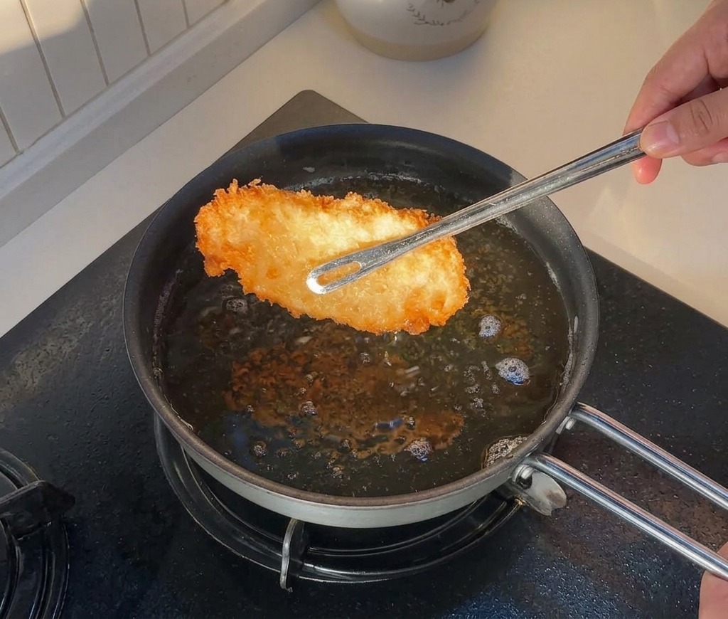 Pinzas levantando un trozo de pollo frito dorado de una sartén con aceite caliente.