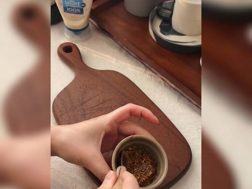 Manos usando una cuchara pequeña para mezclar mostaza en grano y miel en un bol de cerámica.