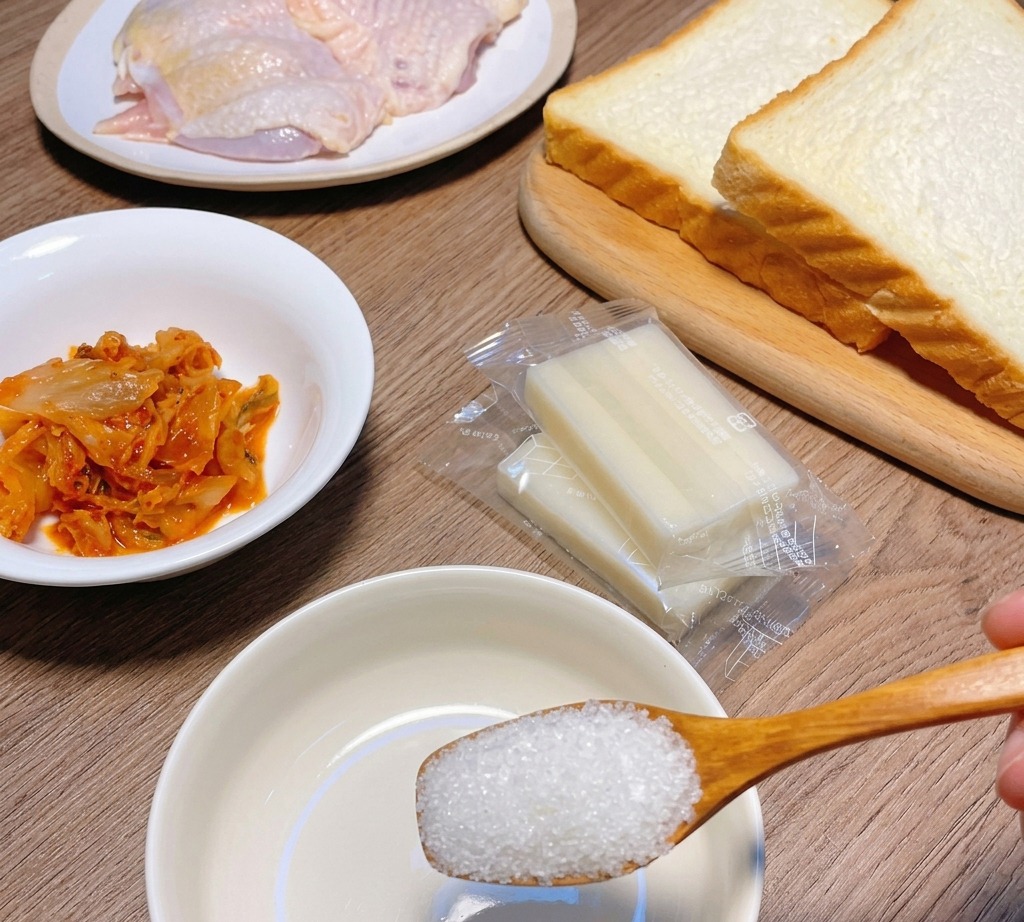 Una cuchara de madera vertiendo azúcar blanca en un pequeño bol blanco rodeado de ingredientes para el sándwich.