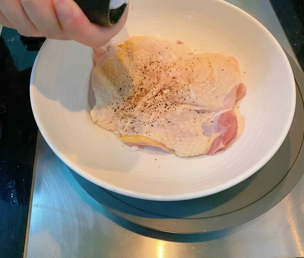 Una mano usando un molinillo de pimienta para sazonar un muslo de pollo crudo en un bol de cerámica.