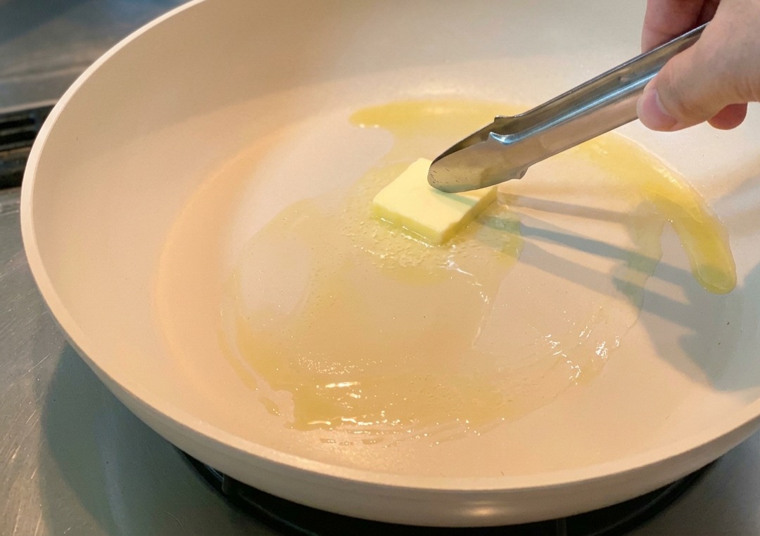 Un trozo de mantequilla derritiéndose en una sartén mientras se mueve con unas pinzas metálicas.