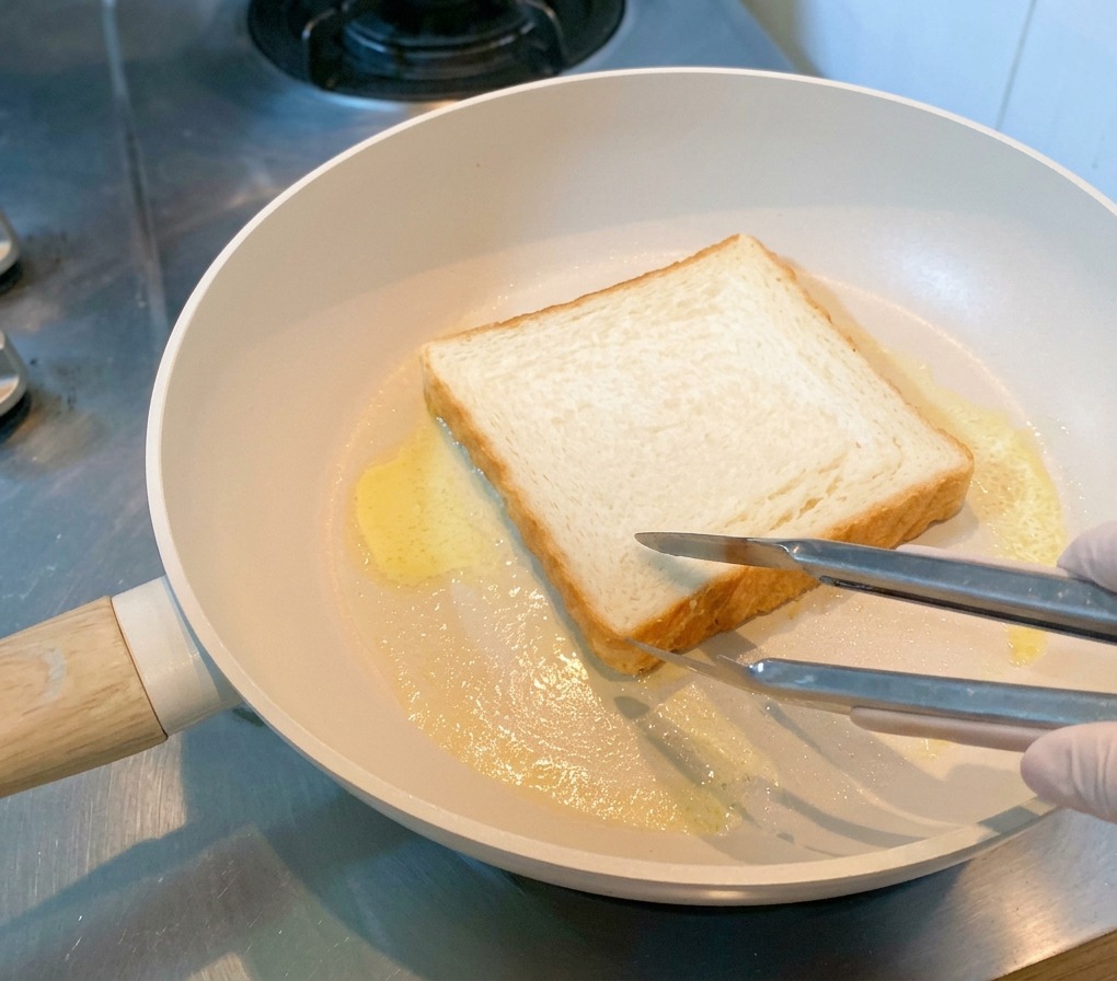Una rebanada de pan blanco tostándose en una sartén con mantequilla derretida, sostenida por unas pinzas.