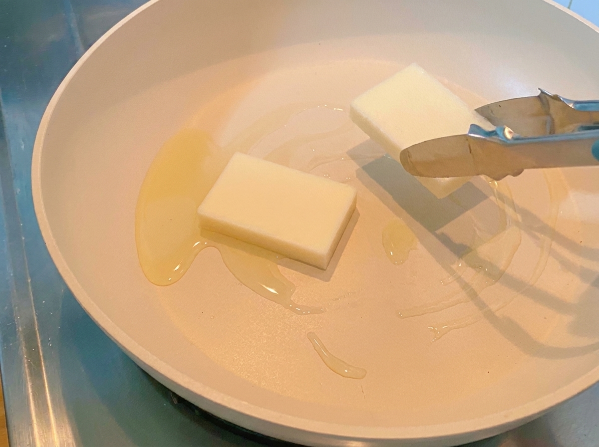 Pinzas colocando dos piezas gruesas y rectangulares de pastel de arroz blanco en una sartén caliente.