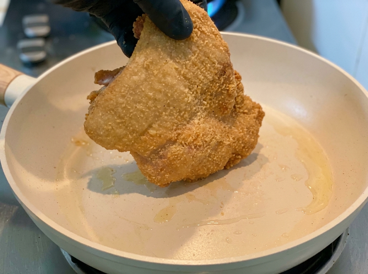 Una mano con guante colocando el pollo rebozado en maicena en una sartén caliente.