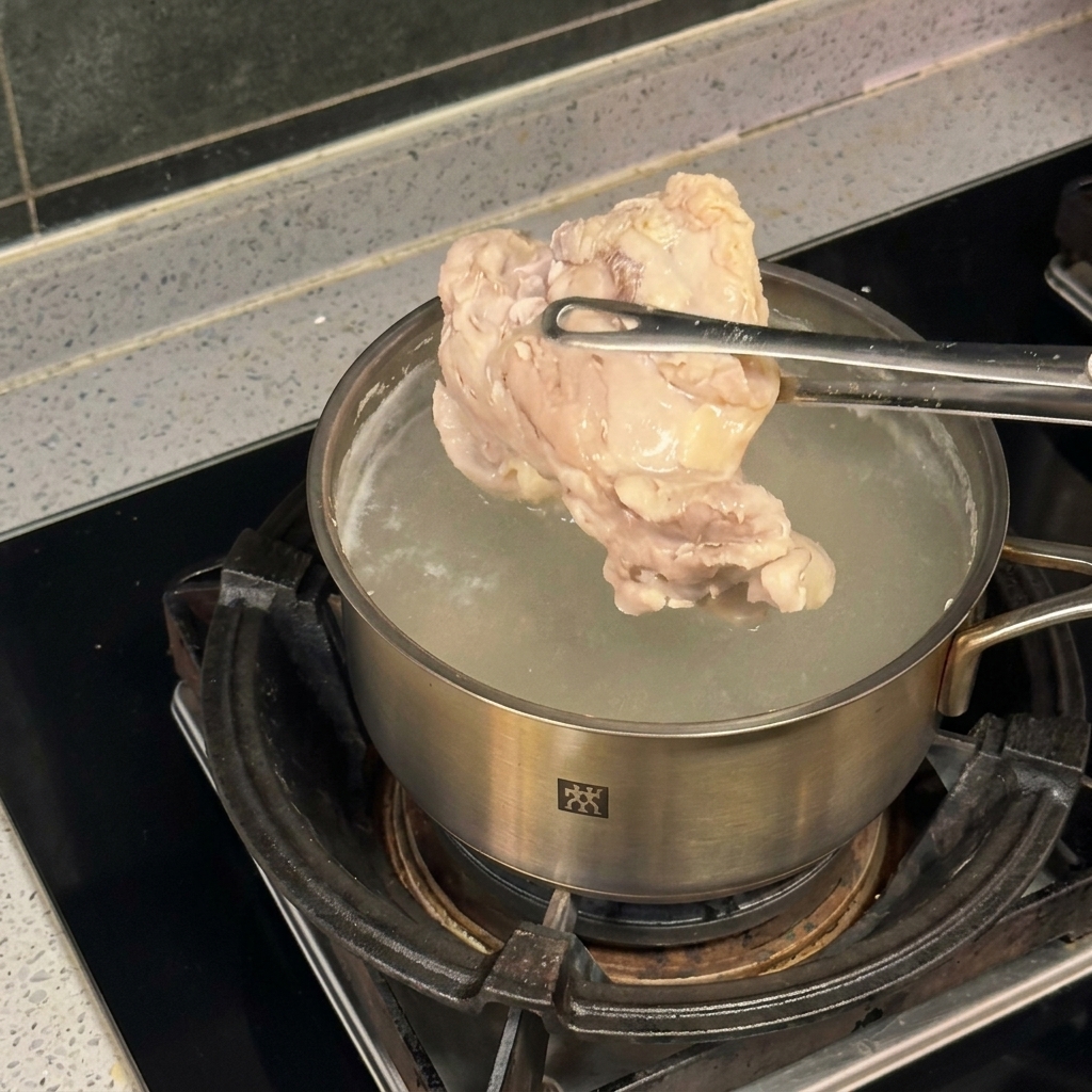 Un trozo de pollo sostenido con pinzas metálicas sobre una olla de acero inoxidable con agua hirviendo.