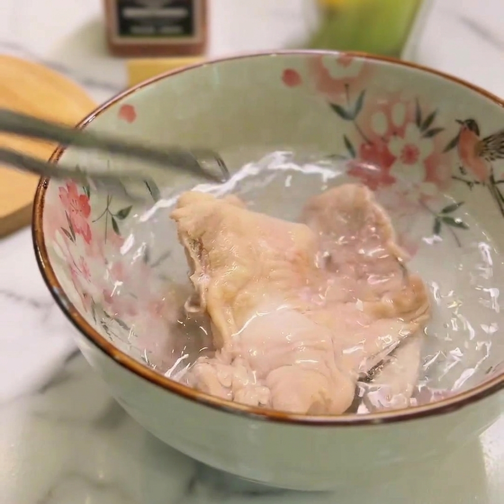 Un trozo de pollo cocido sumergido en un bol de cerámica con agua fría, sujeto brevemente con pinzas.
