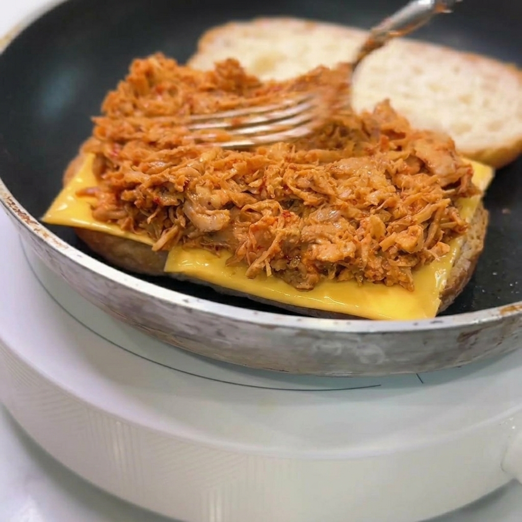 Un tenedor extendiendo una capa generosa de pollo desmechado con salsa naranja sobre el queso en el pan tostado.