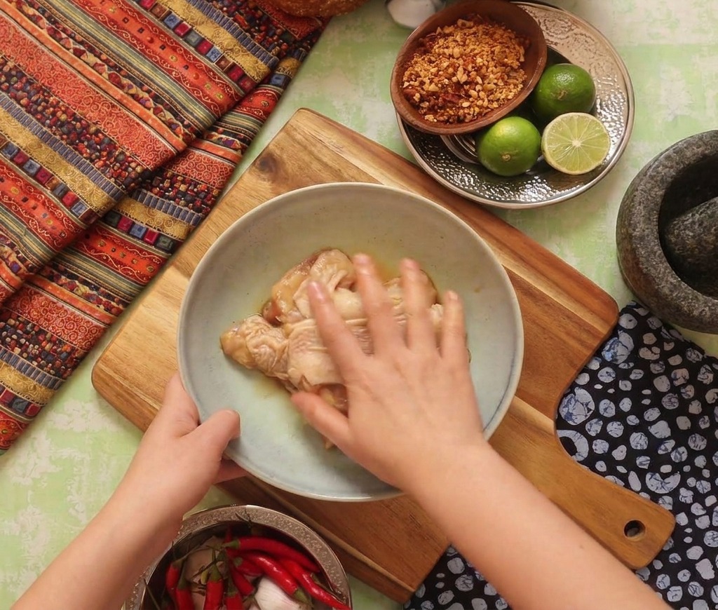 Una mano masajeando un marinado oscuro en los contramuslos de pollo dentro de un bol sobre una tabla.