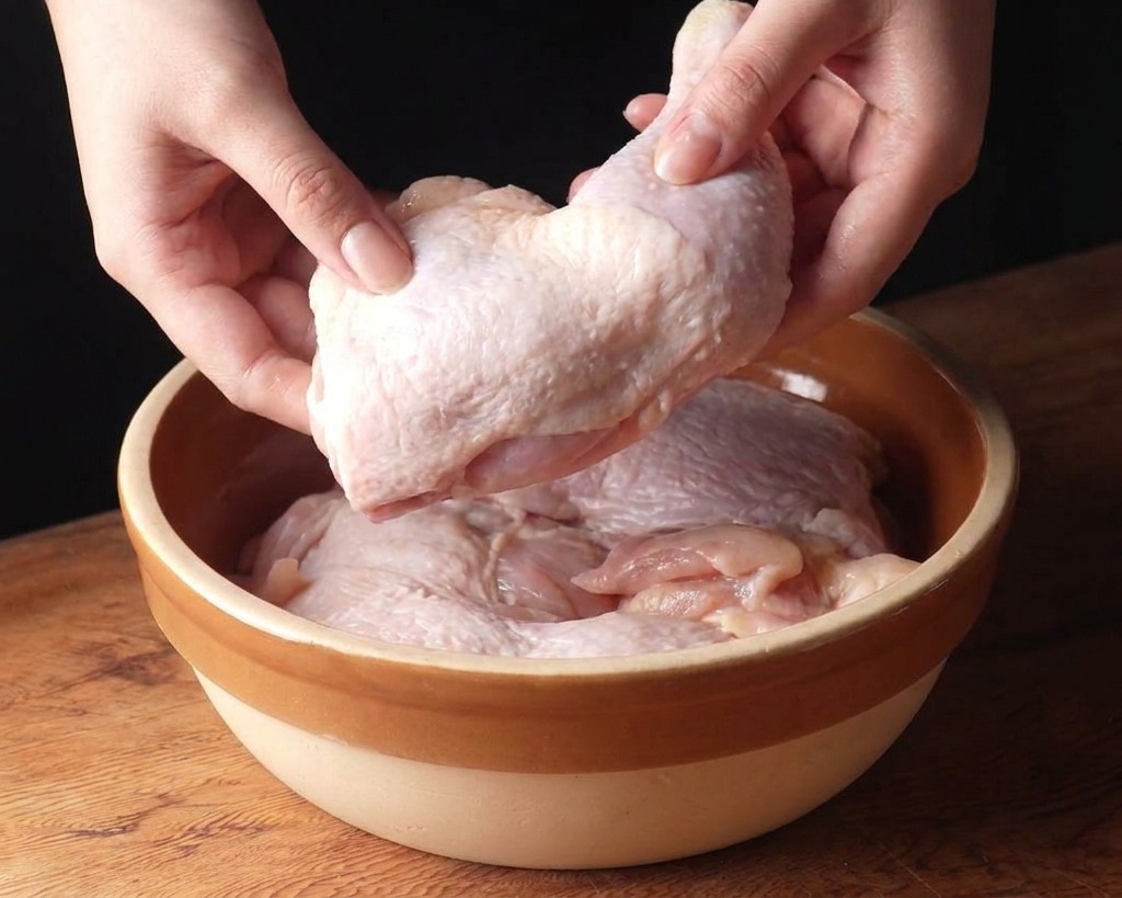 Un par de manos colocando muslos de pollo crudos en un bol de cerámica sobre una superficie de madera.