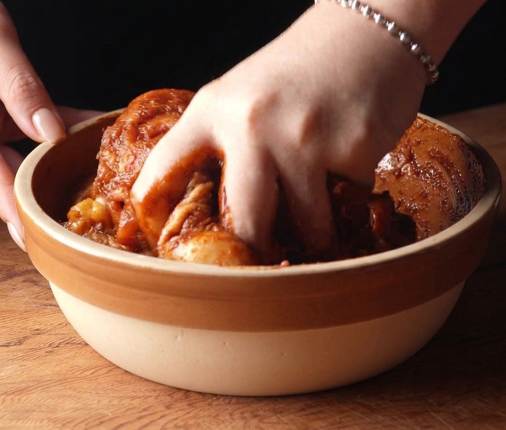 Una mano mezclando muslos de pollo cubiertos en un marinado rojo picante dentro de un bol.