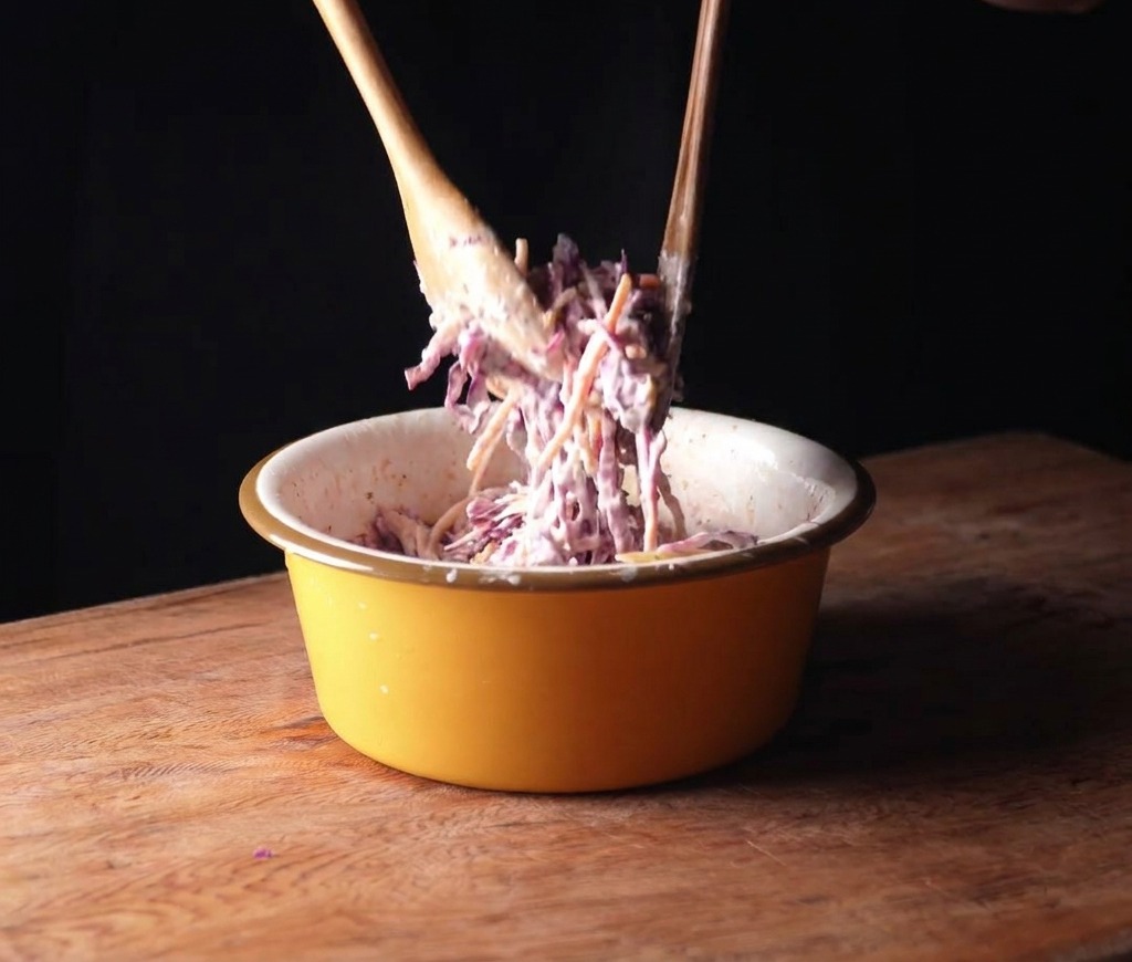 Mezclando repollo morado y zanahorias con aderezo cremoso en un bol amarillo usando cucharas de madera.