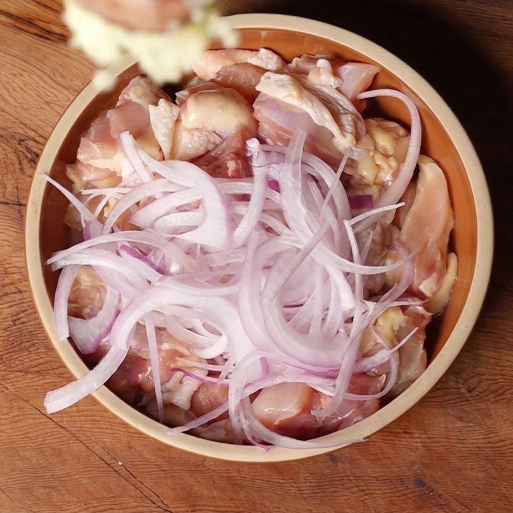 Ajo picado cayendo en un bol de cerámica que contiene trozos de pollo crudo y rodajas de cebolla roja.