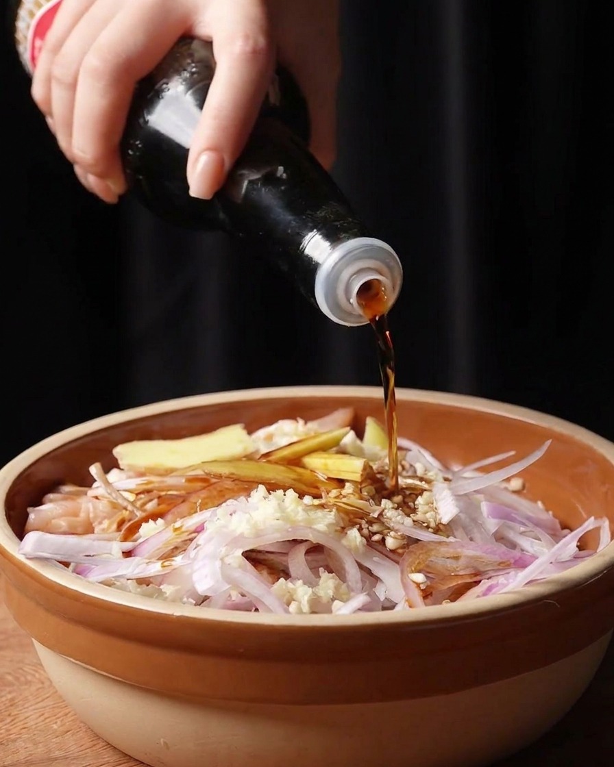 Una mano vertiendo salsa de soja oscura y salsa de pescado desde botellas sobre el pollo y los aromáticos en el bol.