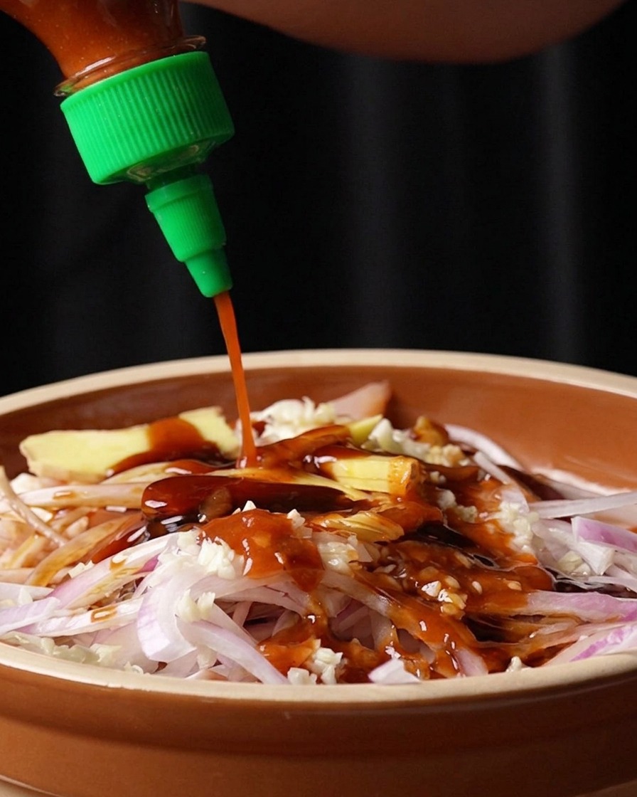 Salsa picante Sriracha roja saliendo de una botella con tapón verde sobre el bol con pollo y especias.