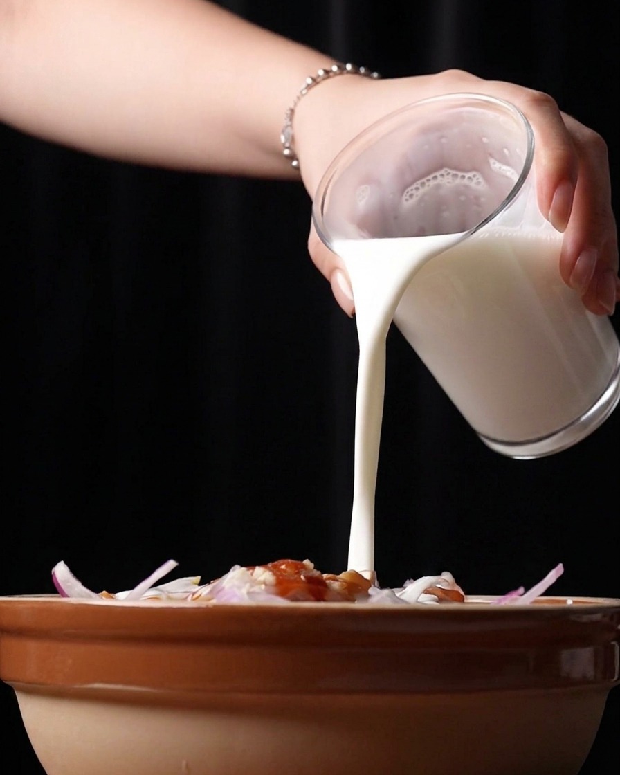 Vertiendo suero de leche blanco y espeso desde un vaso al bol con el pollo troceado y los condimentos.
