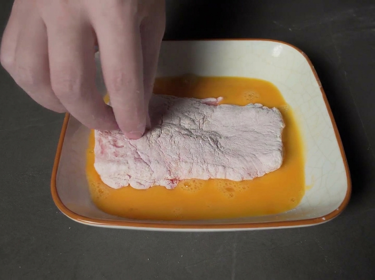 Una mano presionando la chuleta con huevo sobre un cuenco lleno de pan rallado panko.
