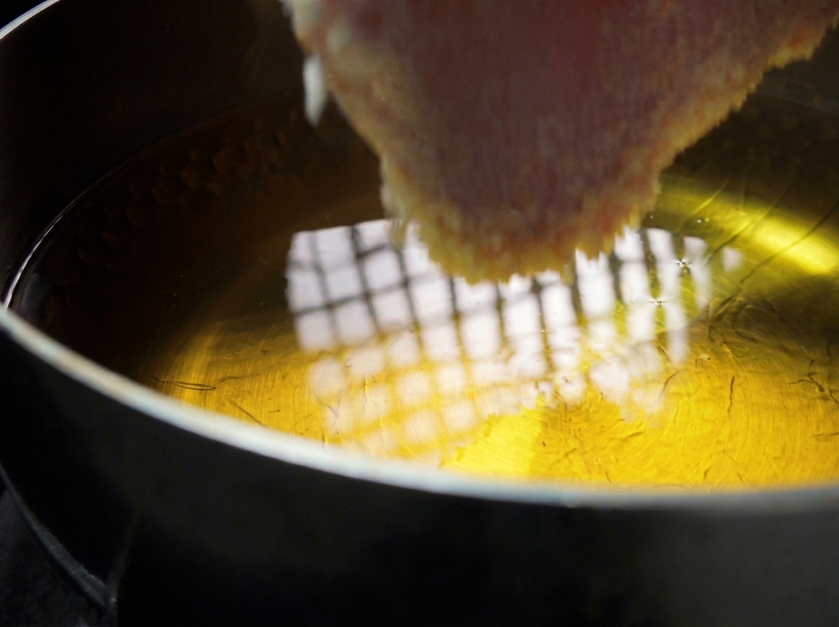 Una chuleta de cerdo empanada siendo introducida con cuidado en una olla con aceite caliente brillante.
