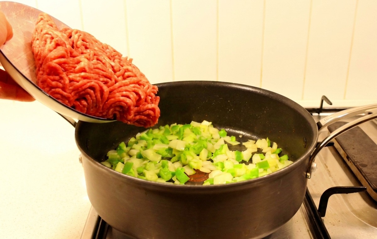 Carne de res molida cruda vertiéndose desde un plato a una sartén que contiene pimientos verdes y cebollas salteadas.