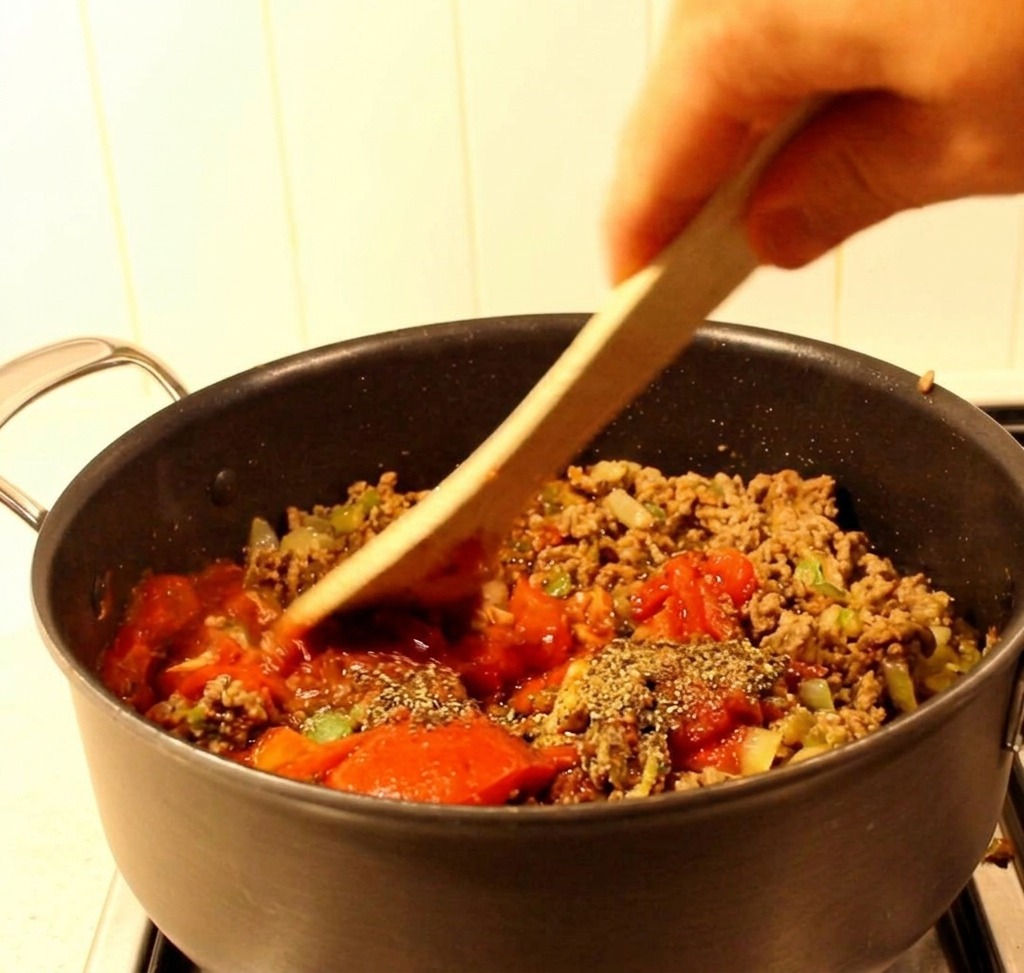 Una espátula de madera mezclando carne molida, pimientos verdes y una salsa de tomate roja en una cacerola negra.