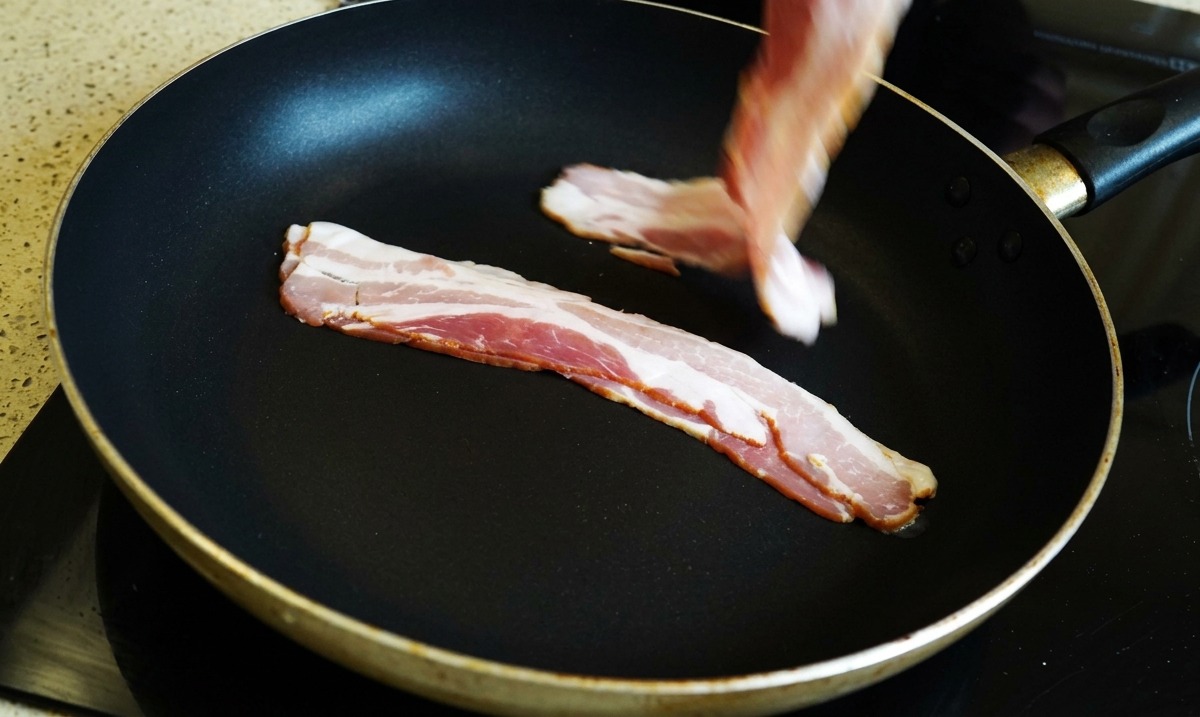 Una mano colocando tiras de tocino crudo en una sartén negra.
