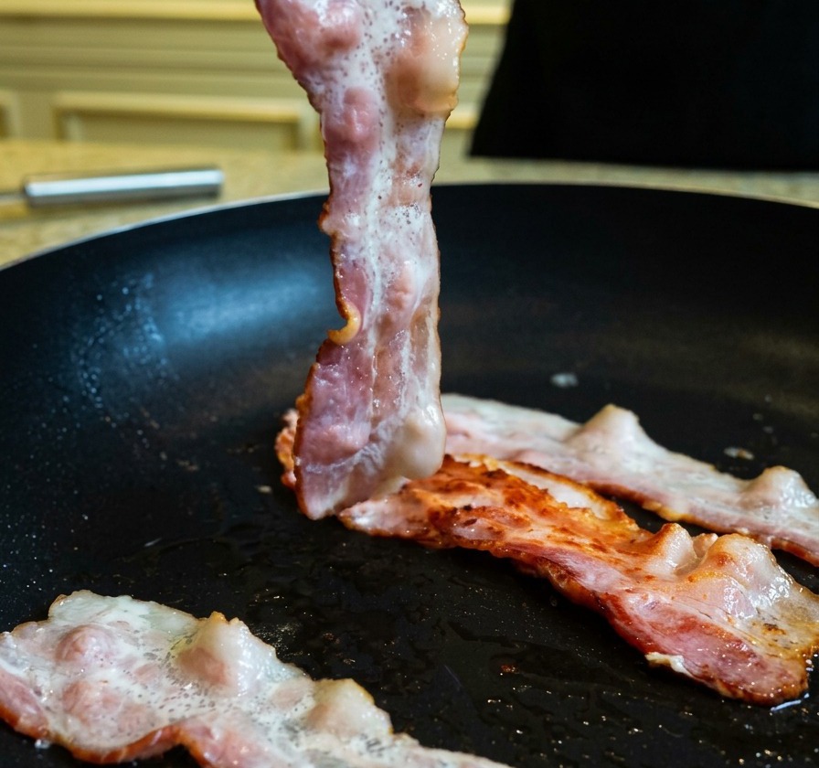 Levantando una tira de tocino parcialmente cocinada y dorada en una sartén usando unas pinzas de cocina.