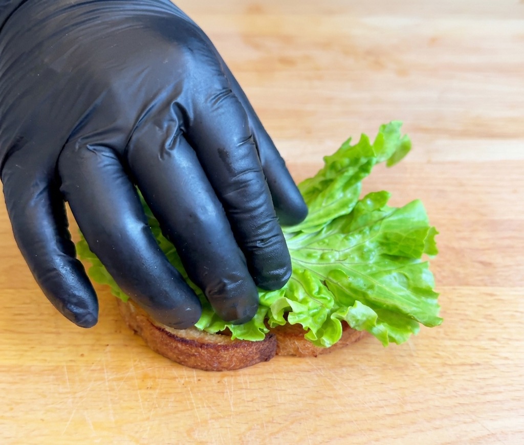 Una mano con guante colocando un trozo de lechuga verde brillante doblada sobre una rebanada de pan tostado que descansa en una tabla de madera.