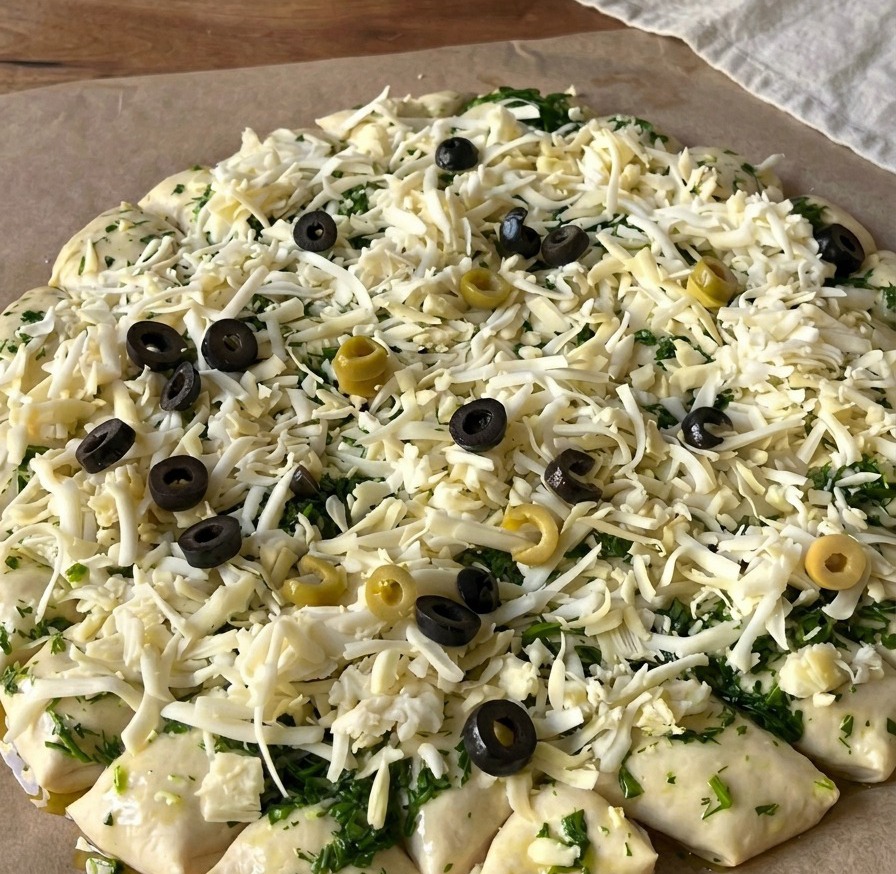 Vista cenital de una masa de pizza cruda cubierta de queso rallado y coronada con numerosas rodajas de aceitunas negras y verdes.