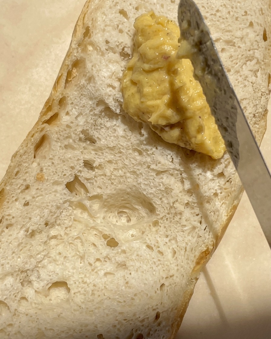 Primer plano de un cuchillo untando una mezcla espesa y cremosa de mantequilla y ajo asado sobre la superficie de una baguette cortada por la mitad.
