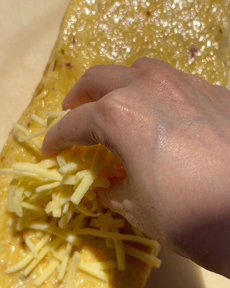 Una mano espolvoreando un gran puñado de queso mozzarella blanco rallado sobre una barra de pan untada con mantequilla de ajo.