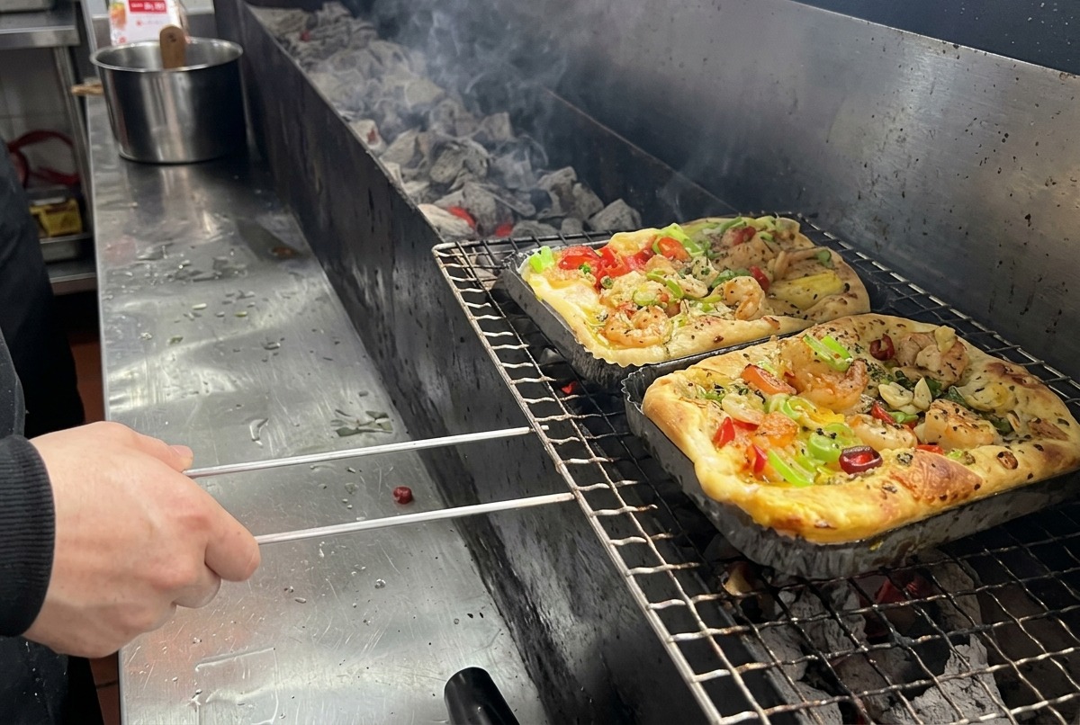 Una pizza rectangular cubierta con camarones y vegetales mixtos colocada sobre una rejilla larga de parrilla de carbón en una cocina profesional.