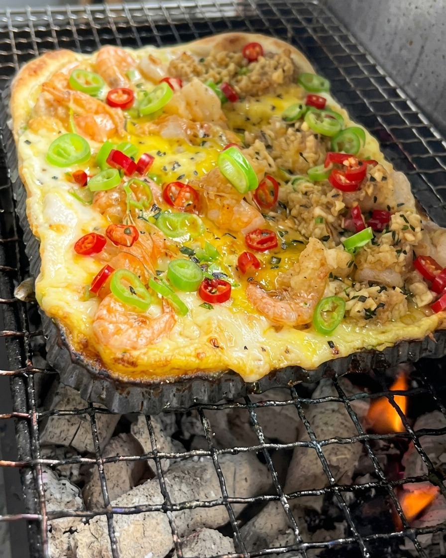 Una masa de pizza rectangular con camarones y vegetales cocinándose sobre brasas de carbón incandescentes.
