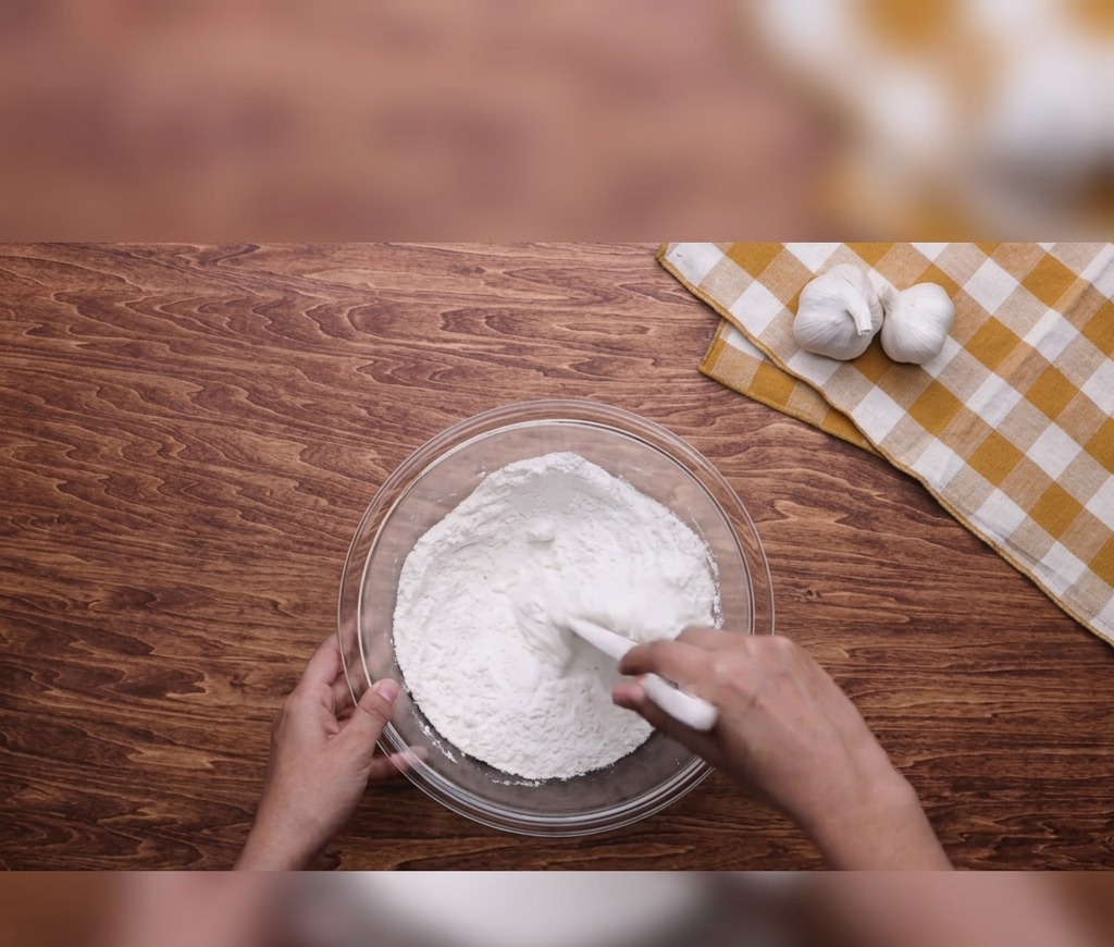 Unas manos usando una espátula blanca para mezclar harina, agua y aceite en un bol de vidrio colocado sobre una mesa de madera.