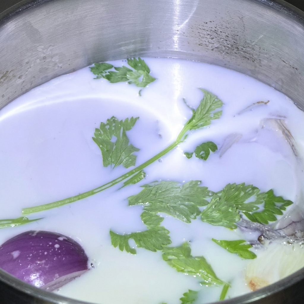 Hierbas verdes frescas y un trozo de cebolla flotando en una olla de acero inoxidable llena de leche blanca.