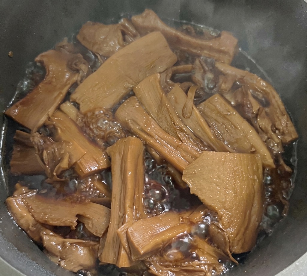 Brotes de bambú en rodajas cocinándose en un líquido de condimento oscuro y burbujeante dentro de una olla metálica.