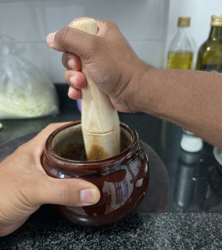 Alguien machacando ajo frito oscuro con una mano de mortero de madera dentro de un pequeño cuenco de cerámica.