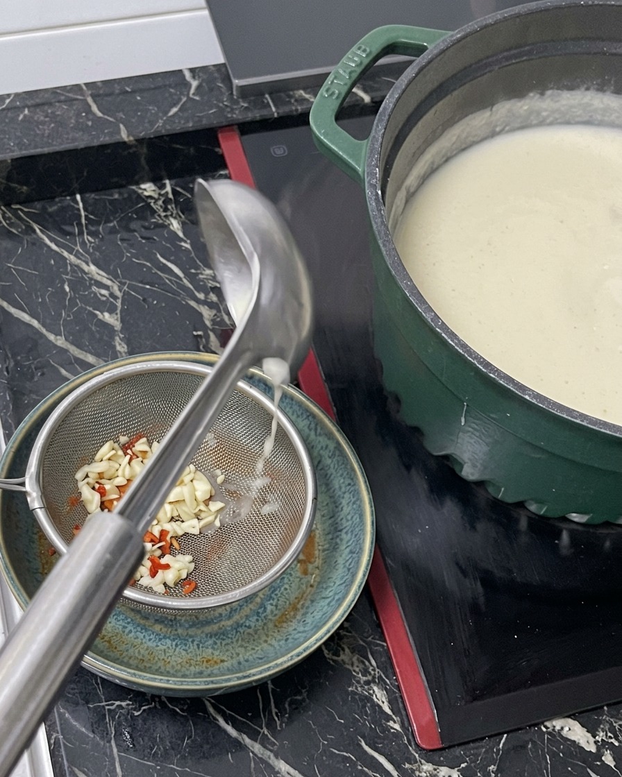 Caldo blanco y muy caliente vertiéndose desde un cazo a través de un colador metálico en un tazón con la base de condimento.