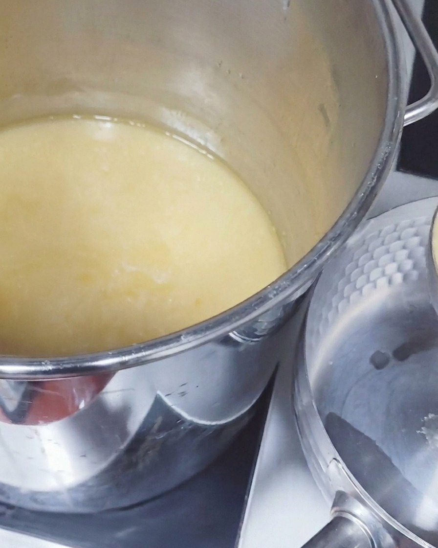 Un cucharón de metal grande sacando caldo de cerdo opaco y de color amarillo pálido de una olla profunda para verterlo en un cazo más pequeño.