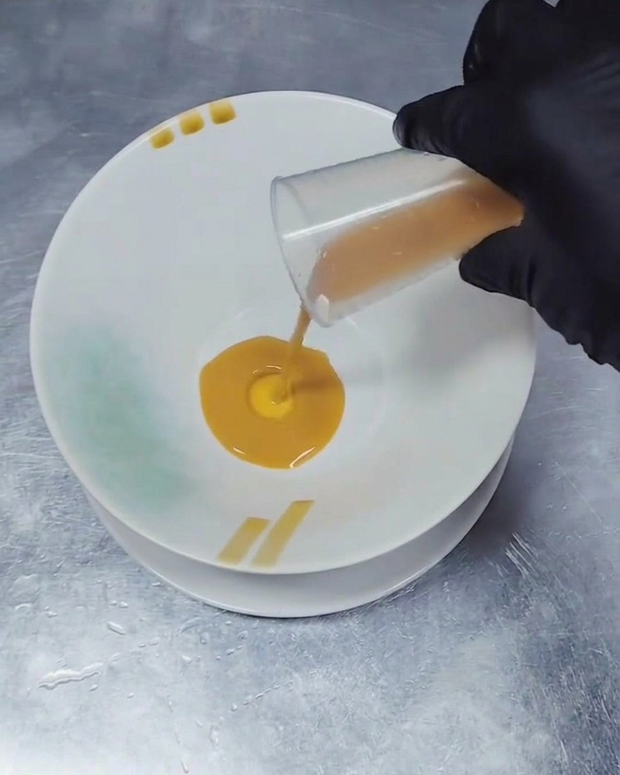 Una mano con guante vertiendo una pequeña cantidad de condimento líquido de color marrón oscuro desde un vaso medidor transparente hacia el centro de un tazón de cerámica blanca.