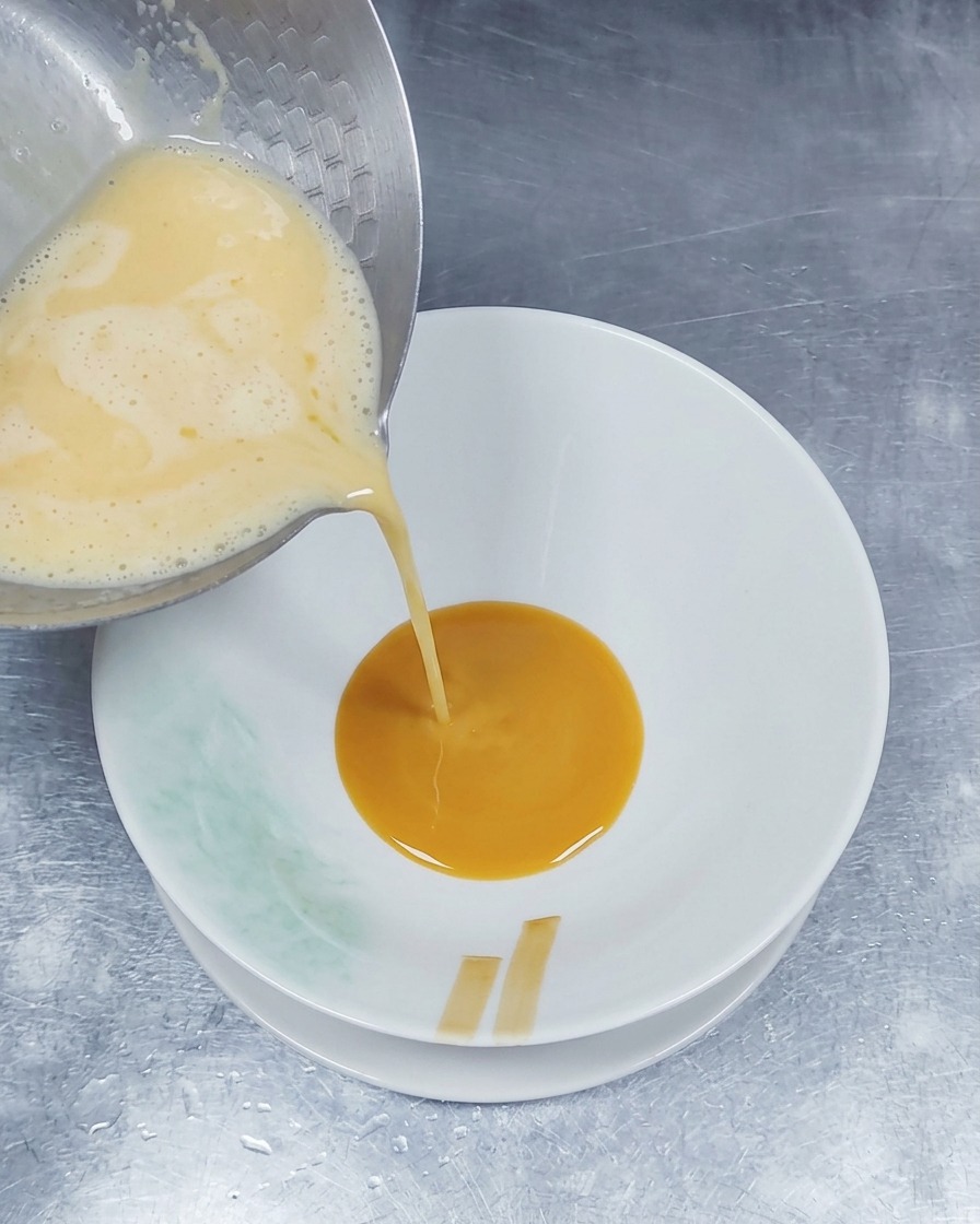 Caldo de cerdo caliente y espumoso vertiéndose desde un cazo de metal en un tazón que ya cuenta con una capa de salsa de condimento marrón.