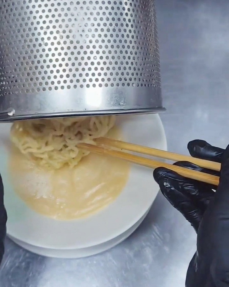 Un colador de fideos de acero inoxidable deslizando fideos ramen ondulados dentro de un tazón con caldo cremoso Tonkotsu.