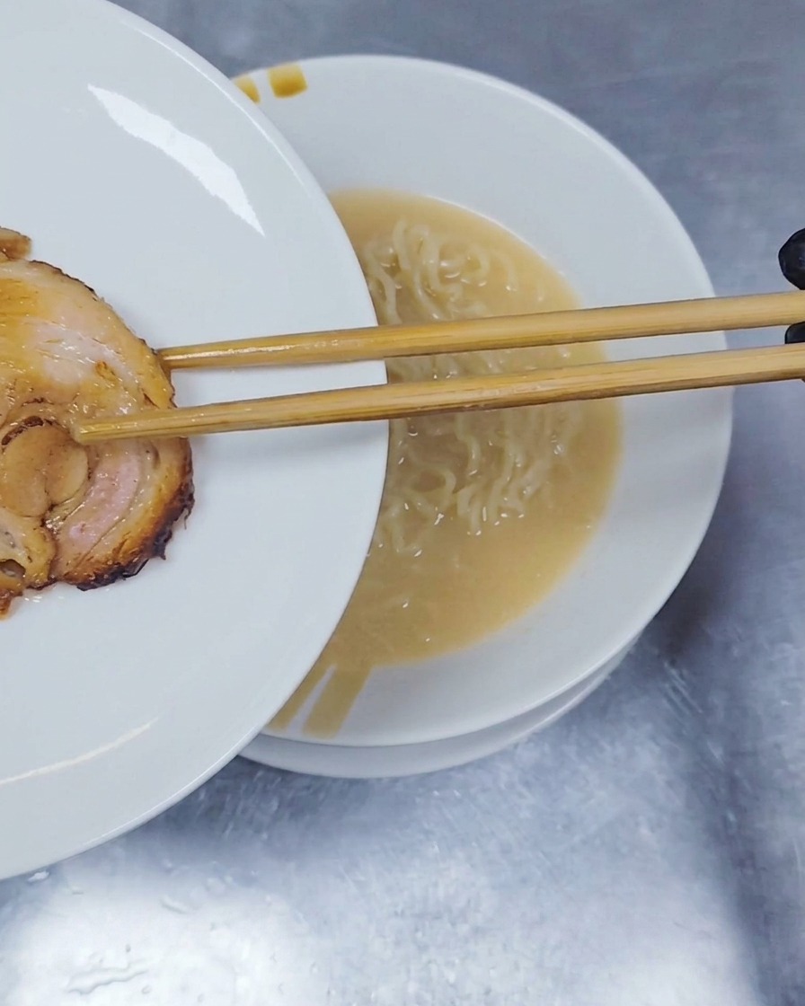 Una rodaja de cerdo chashu enrollado y estofado siendo colocada encima de los fideos ramen con unos palillos de madera.