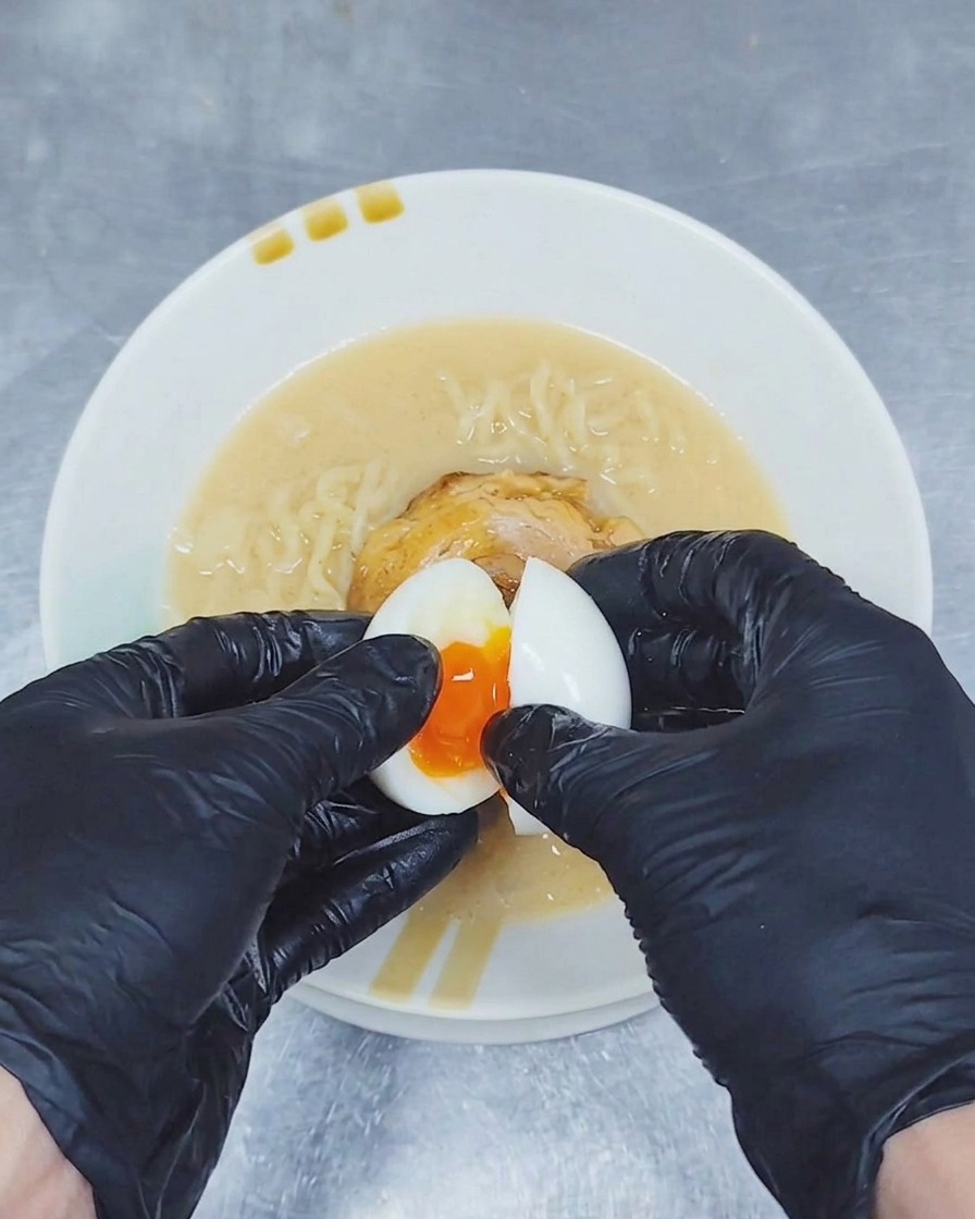 Un par de manos con guantes partiendo un huevo cocido sobre un tazón de ramen para mostrar una yema dorada y líquida.