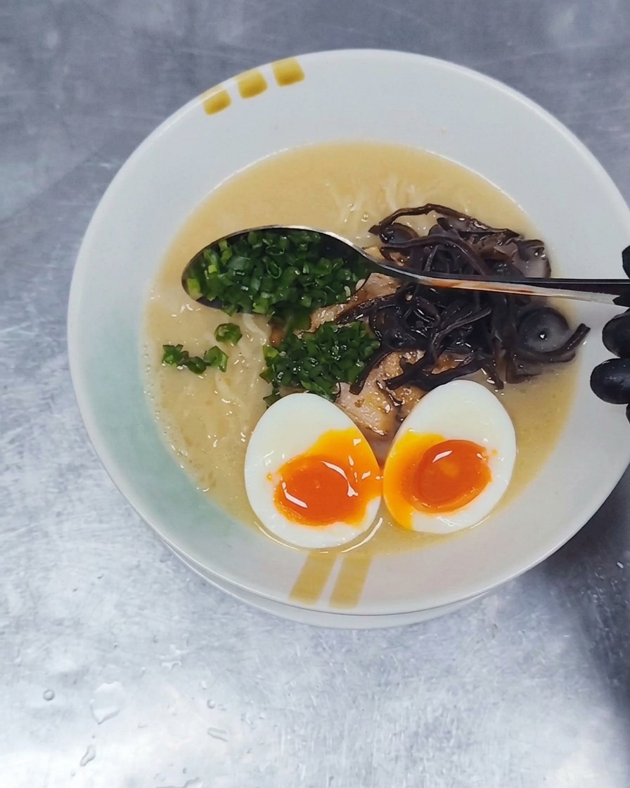 Una cuchara de metal esparciendo cebollín picado sobre la superficie de un tazón de ramen Tonkotsu.