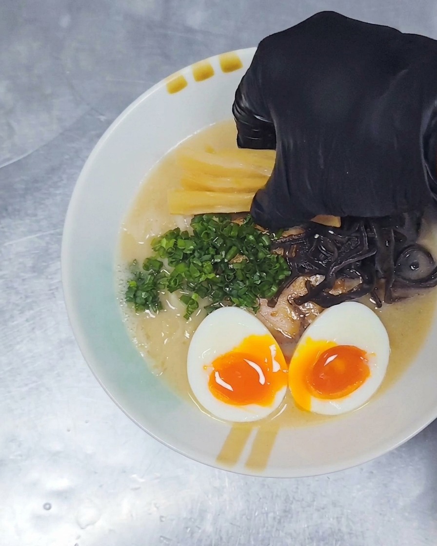 Una mano con guante colocando tiras amarillas de brotes de bambú sazonados en un tazón de ramen que ya tiene cerdo, huevo y cebollín.