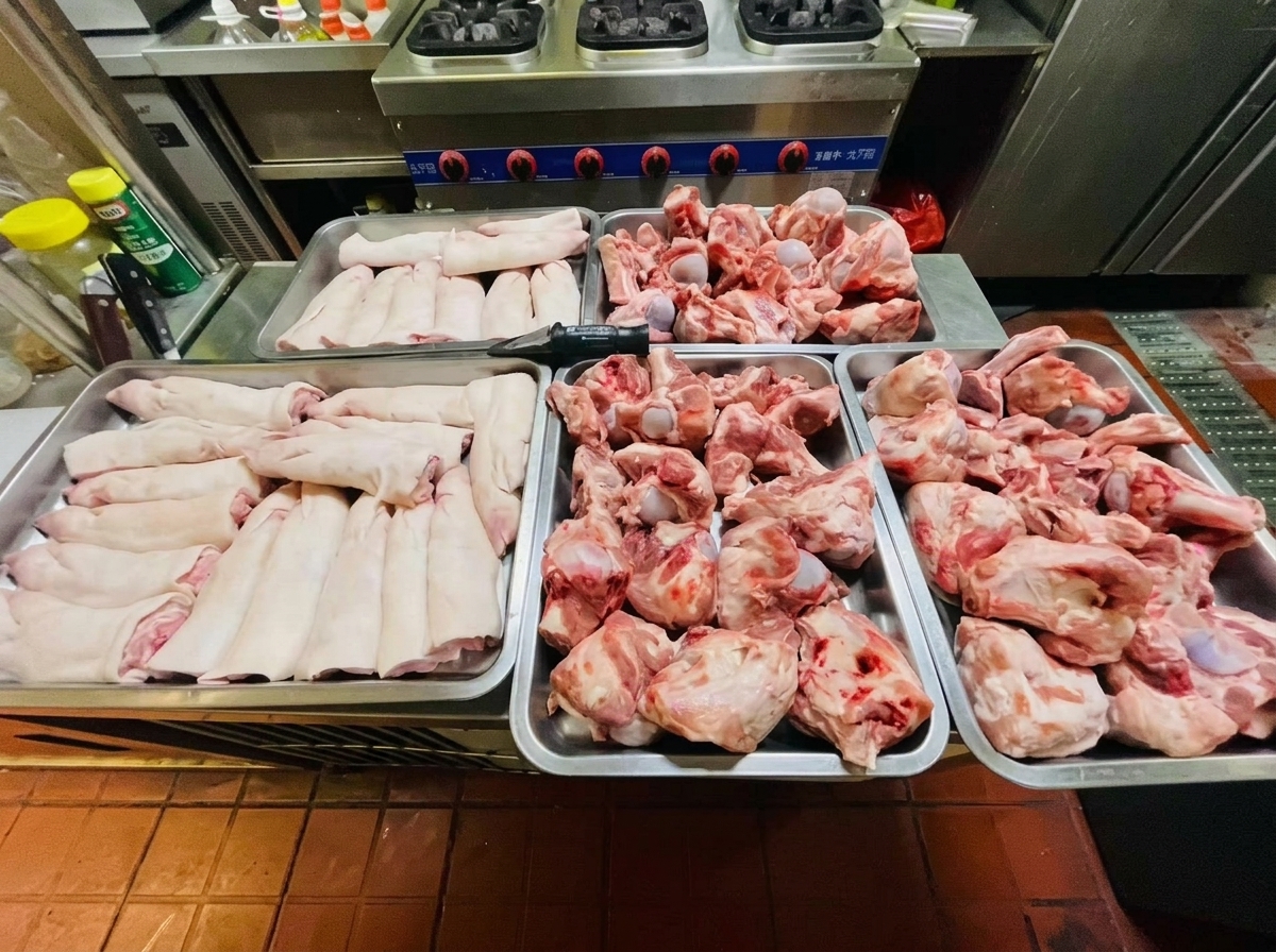 Cuatro bandejas metálicas llenas de huesos de codillo y manitas de cerdo crudas sobre una encimera de acero inoxidable.