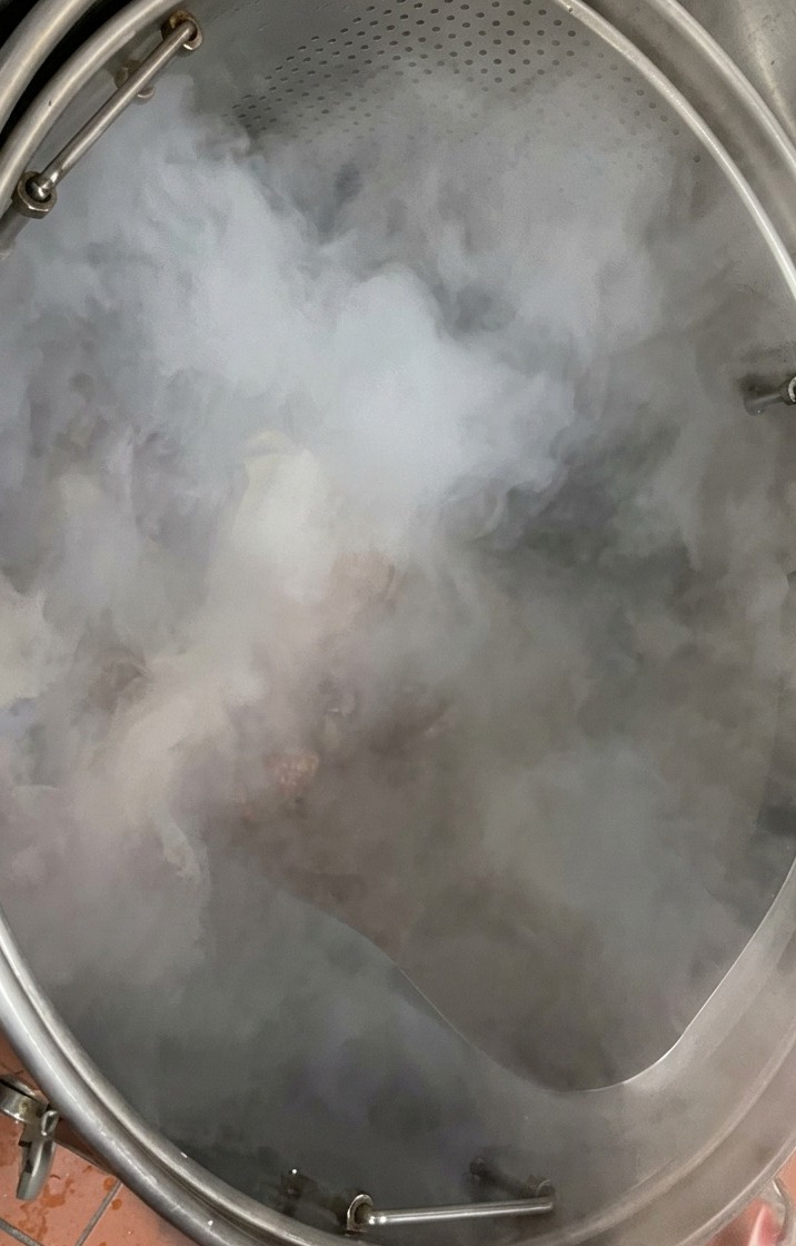 Una bandeja con huesos de cerdo crudos vertiéndose en una gran olla de acero inoxidable con agua hirviendo.