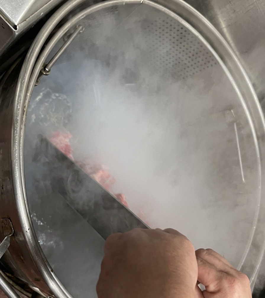 Unas manos sosteniendo una gran paleta metálica para remover los huesos de cerdo dentro de una olla industrial humeante.
