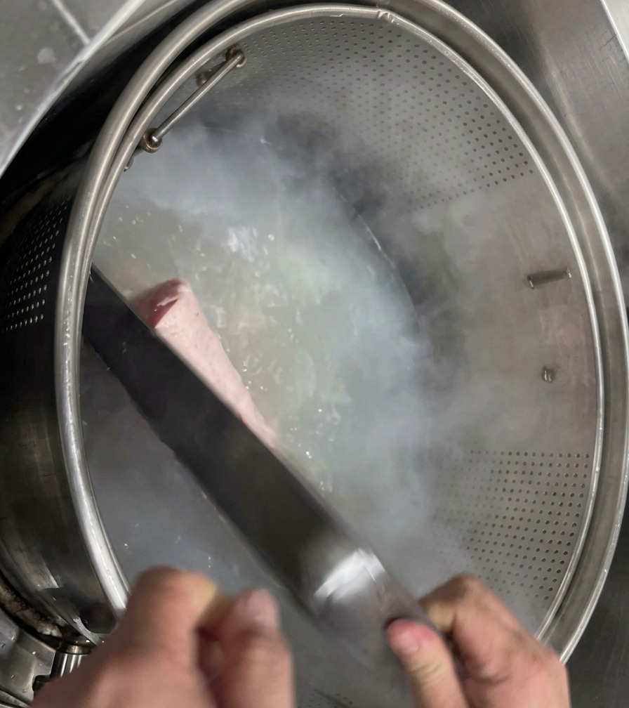 Manitas de cerdo limpias y blanqueadas introduciéndose en una canasta de malla metálica dentro de una gran olla con agua hirviendo limpia.