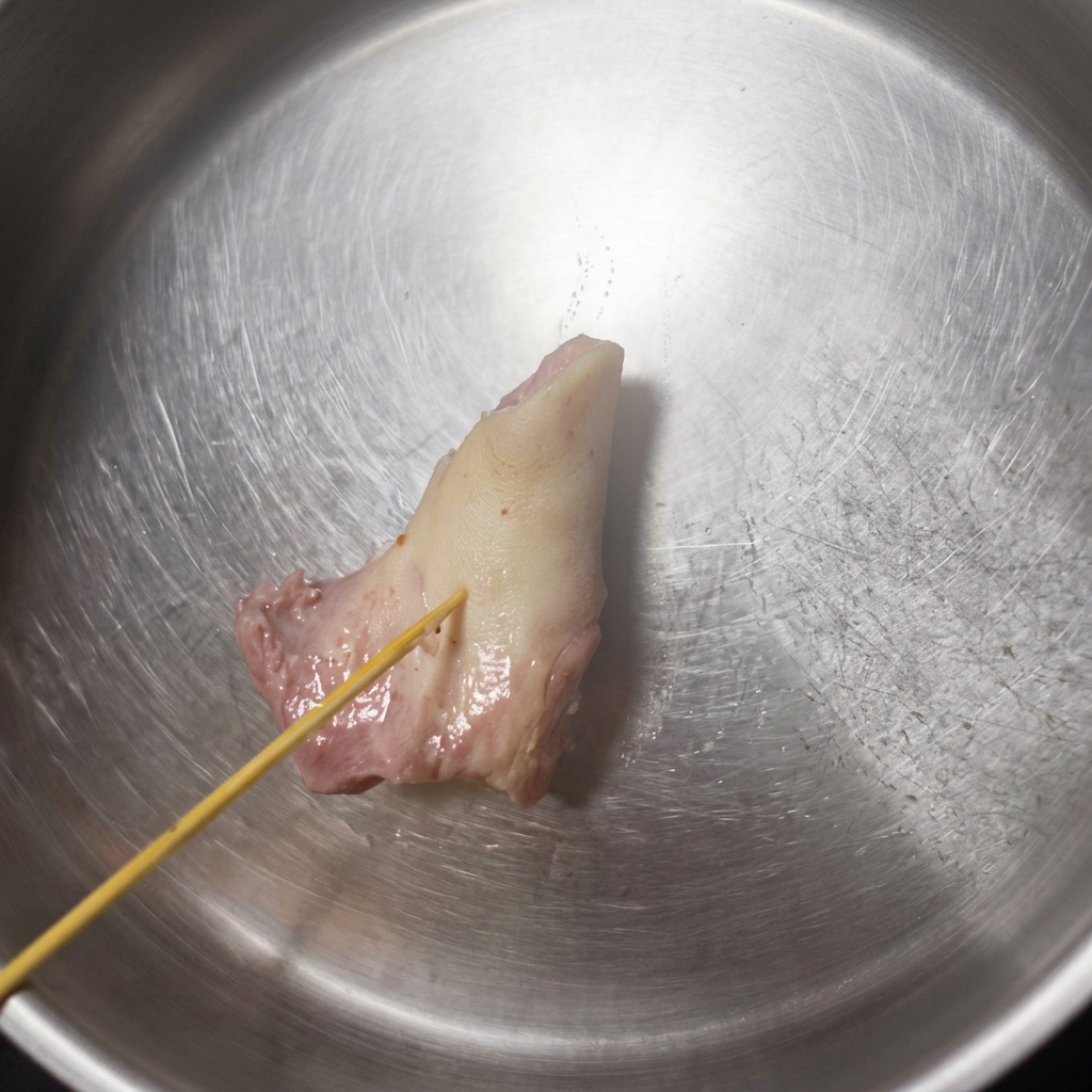 Brocheta de madera presionando un hueso de cerdo ablandado y pálido dentro de un tazón de acero inoxidable, mostrando cómo empieza a ceder.