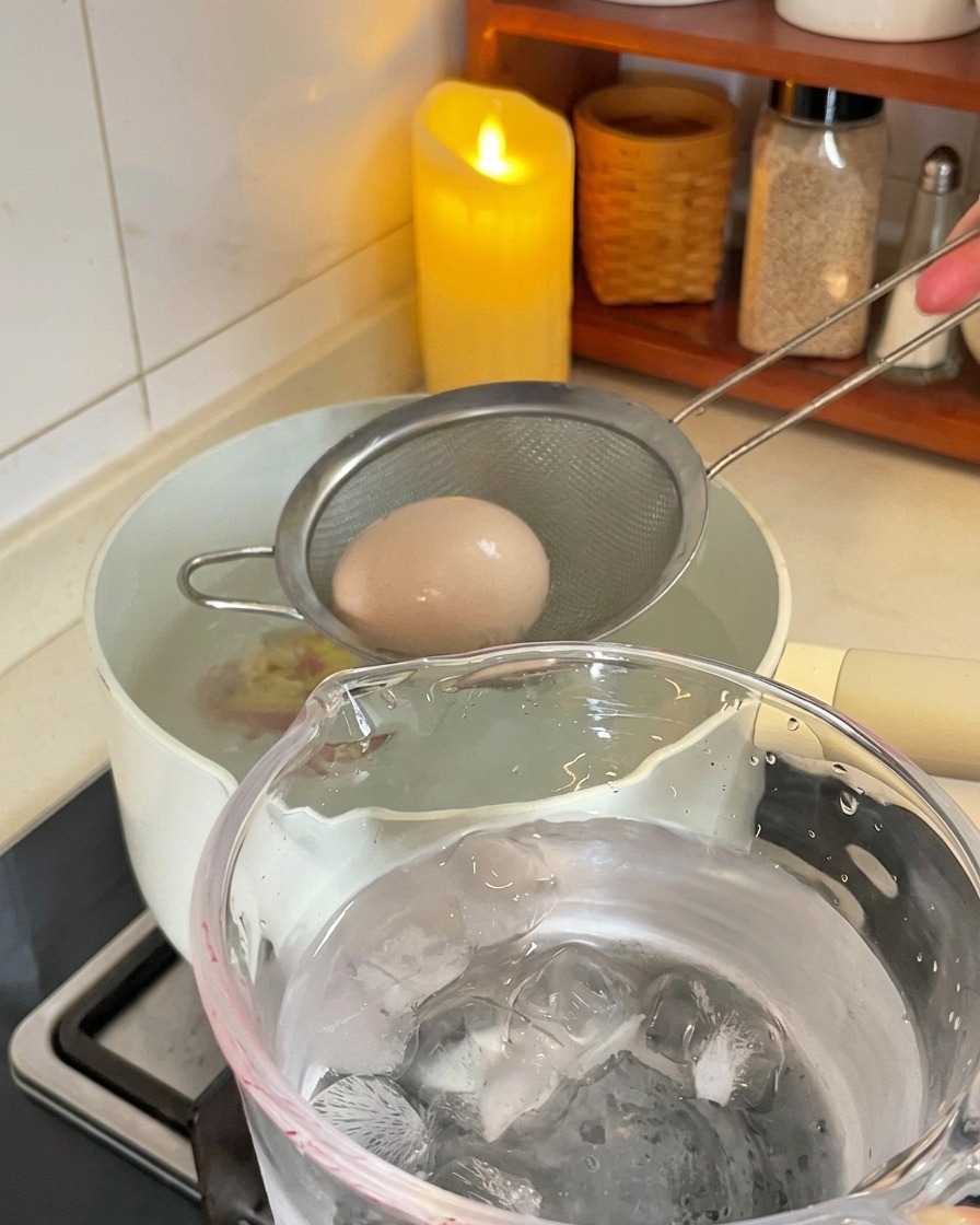 Un colador metálico sacando un huevo cocido de la olla hacia un vaso lleno de agua con hielo.
